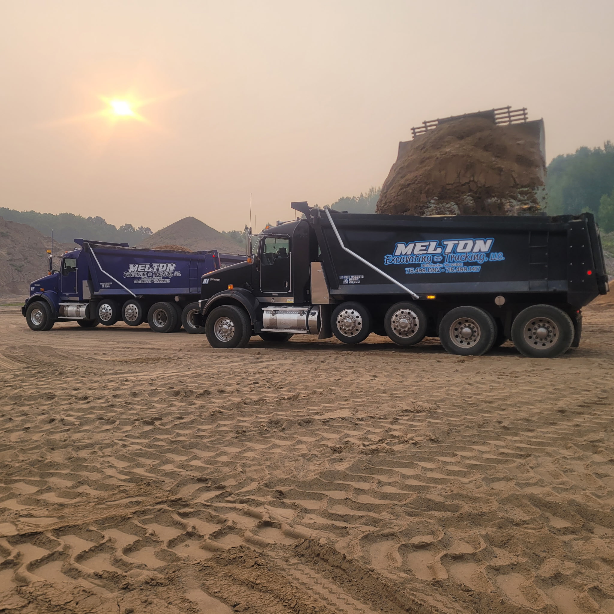 Two Melton Excavating & Trucking dump trucks at a material yard